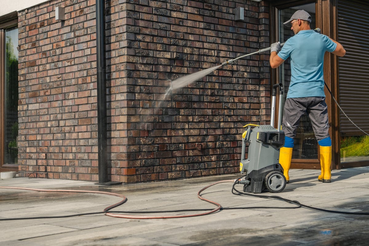 Technician in yellow boots pressure washing a brick wall of a modern house